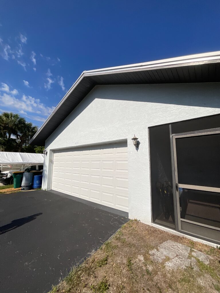 A newly painted white stucco house with a clean white garage door and dark gray soffit under a blue sky.